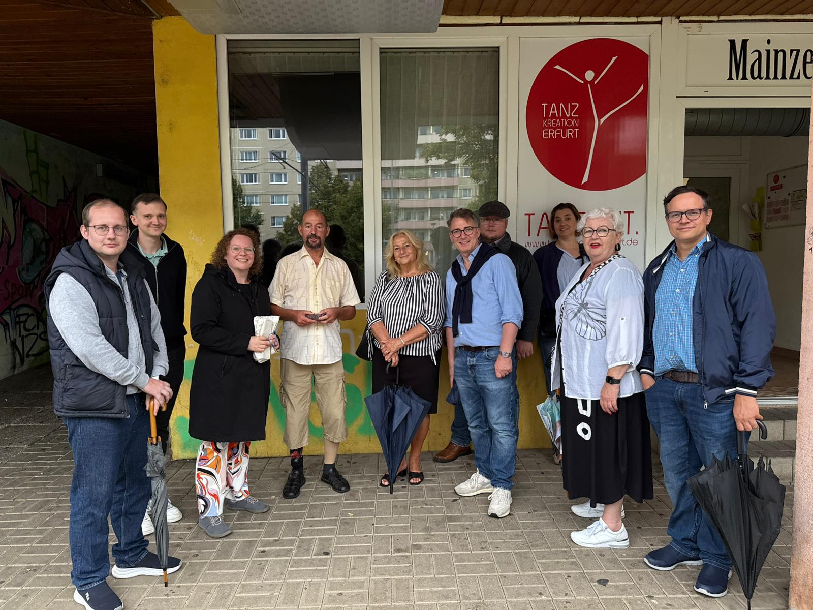 Gruppe von zehn Personen beim Stadtteilspaziergang der FDP Erfurt im Rieth. Sie stehen vor dem Eingang der Tanzschule „Tanz Kreation Erfurt“, lächeln in die Kamera und halten teils Regenschirme in der Hand. Im Hintergrund sind ein gelb gestrichener Gebäudeeingang, Fenster eines Wohnblocks und ein rotes Logo der Tanzschule zu sehen.