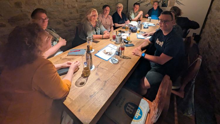 Eine Gruppe von etwa zehn Personen sitzt an einem langen Holztisch in einem historischen Kellerraum mit Steinwänden. Die Stimmung ist freundlich und lebendig. Die Blickrichtung geht über die Schulter einer Teilnehmerin hinweg auf den vollen Tisch. Die Gäste sind im Gespräch vertieft, einige mit Getränken in der Hand. Die Atmosphäre wirkt konzentriert und zugleich entspannt.