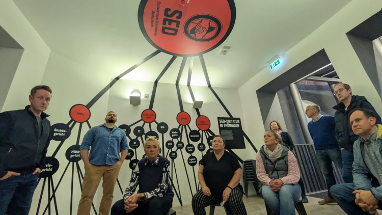 Mehrere Personen sitzen und stehen in einem Ausstellungsraum zur SED-Diktatur in Thüringen. Über ihnen verläuft eine grafische Darstellung der Machtstrukturen der SED, die von einem großen roten Kreis an der Decke mit dem SED-Logo ausgeht und sich mit schwarzen Linien an den Wänden verzweigt. Die Besucher:innen blicken aufmerksam nach oben in Richtung der Darstellung.