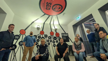 Mehrere Personen sitzen und stehen in einem Ausstellungsraum zur SED-Diktatur in Thüringen. Über ihnen verläuft eine grafische Darstellung der Machtstrukturen der SED, die von einem großen roten Kreis an der Decke mit dem SED-Logo ausgeht und sich mit schwarzen Linien an den Wänden verzweigt. Die Besucher:innen blicken aufmerksam nach oben in Richtung der Darstellung.